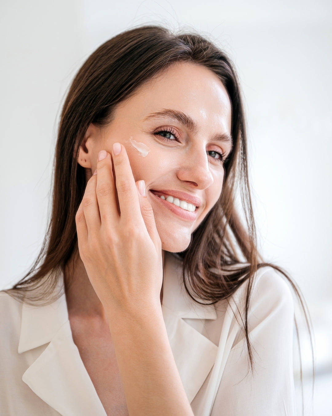 Woman smiling while applying Aminocare® Reverse Face Cream to her cheek for hydrated, radiant winter skin.